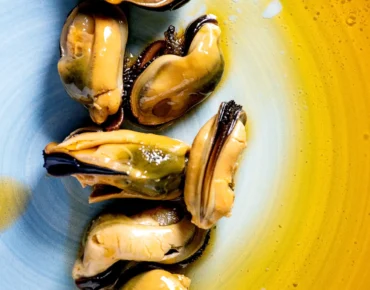 Mejillones frescos en un plato azul con un fondo amarillo, servidos con un toque de aceite de oliva. Ideal para un plato de mariscos gourmet.