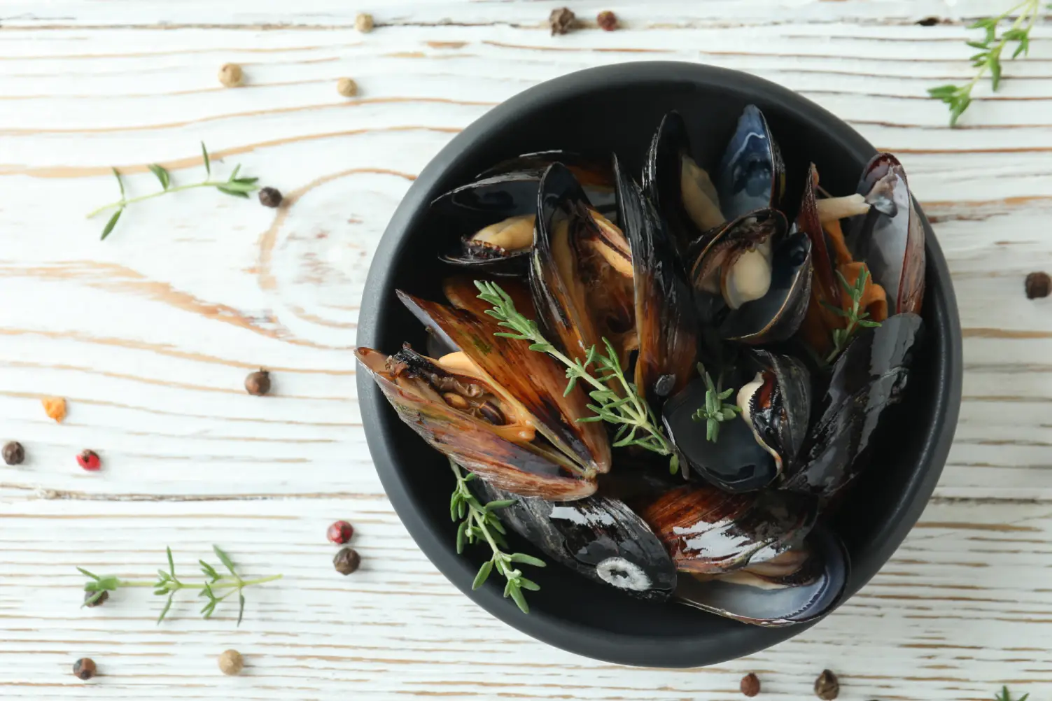 Plato de mejillones frescos en un tazón negro, decorado con hierbas frescas y especias sobre una mesa de madera clara.