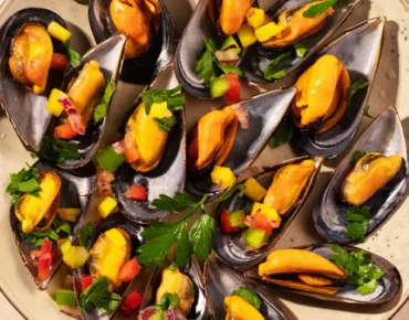 Plato de mejillones al vapor con salsa de mango y pimientos, decorado con perejil fresco. Ideal para una comida gourmet o aperitivo.